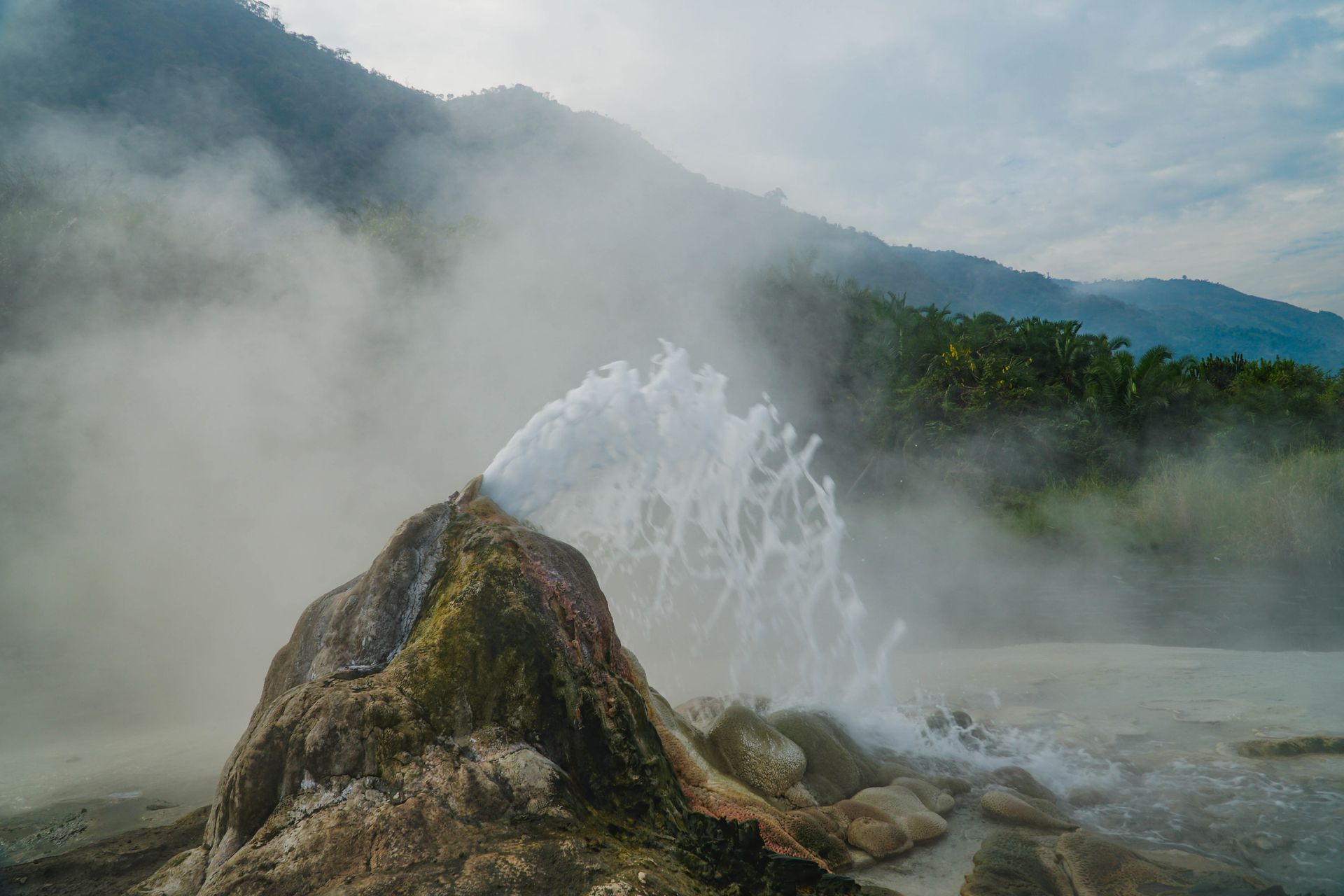 A geothermal geyser erupts with a powerful jet of water and steam from a rocky mound against a backdrop of hazy hills.