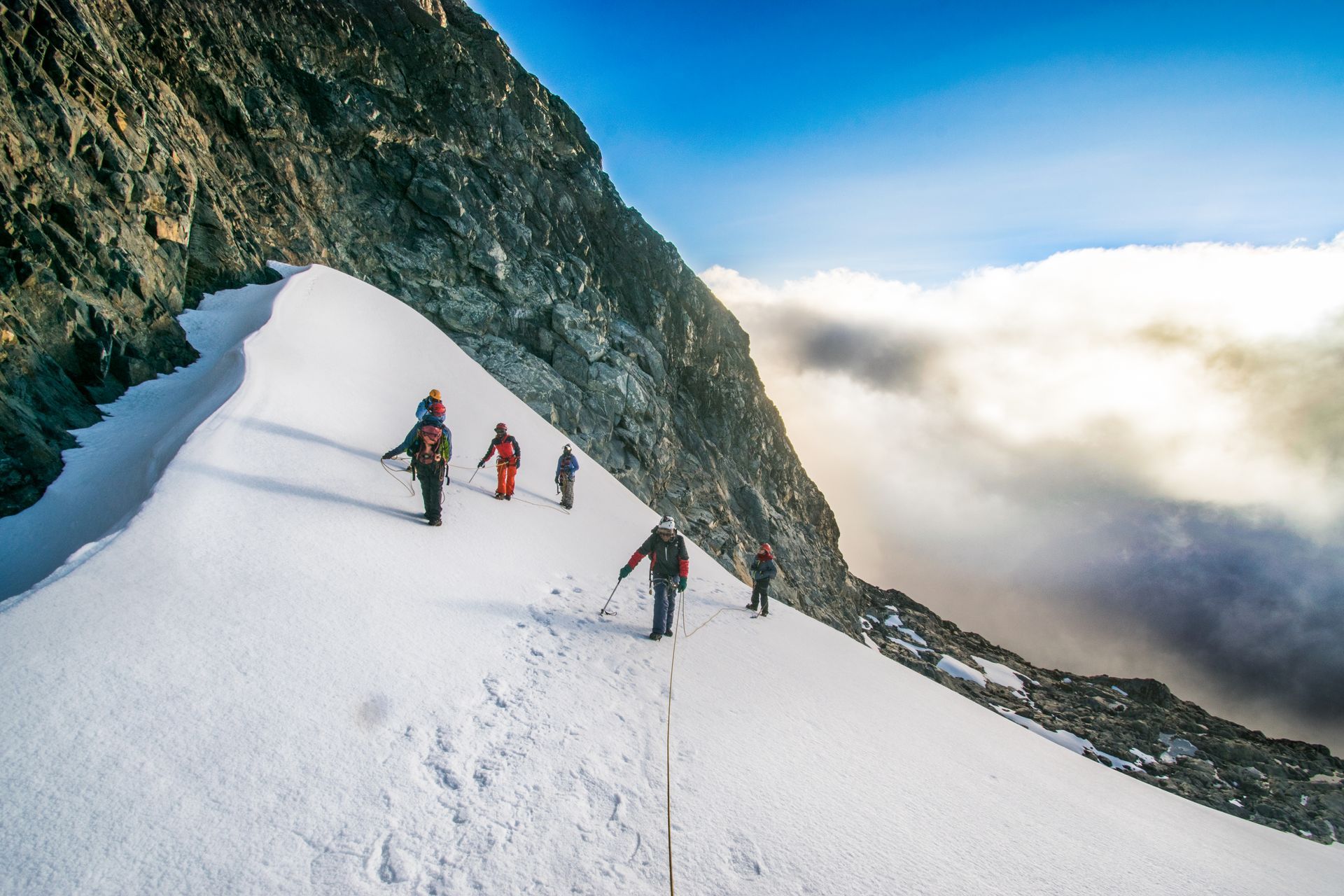 A group of climbers hike across a narrow, snowy mountain ridge alongside a steep rock face under a bright blue sky.