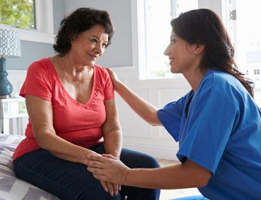 A female nursing kneeling in front of a seated woman hold her hand and shoulder