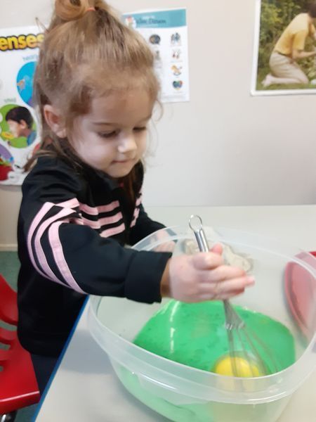 Girl mixing green batter with a whisk in a clear bowl on a table.
