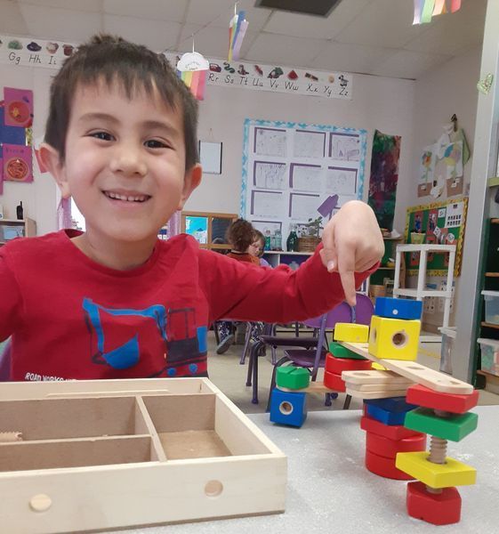 Boy points at colorful building blocks he constructed at a table in a classroom.