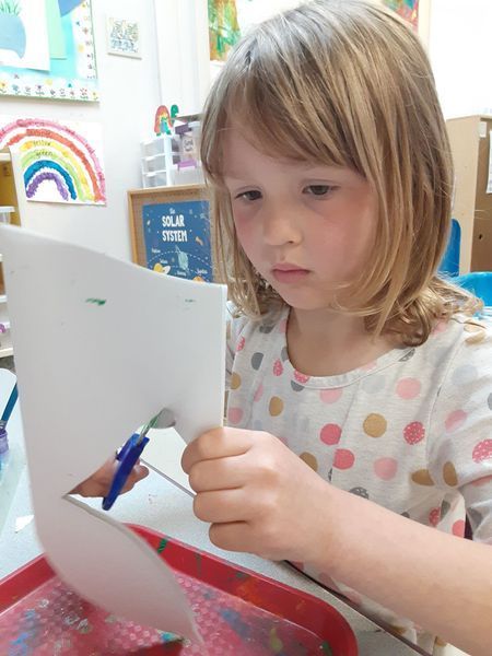 A child with blonde hair cutting white paper with blue scissors in a classroom setting.