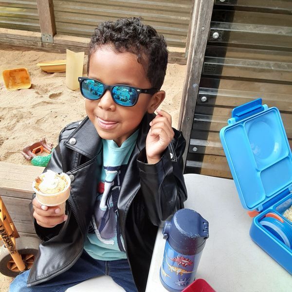 Boy in sunglasses and leather jacket holds ice cream, sitting near a lunchbox and sandbox.