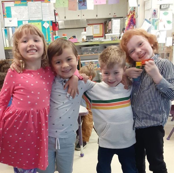 Four children smiling, arms around each other. Classroom setting. Girl in pink dress, others in casual clothing.