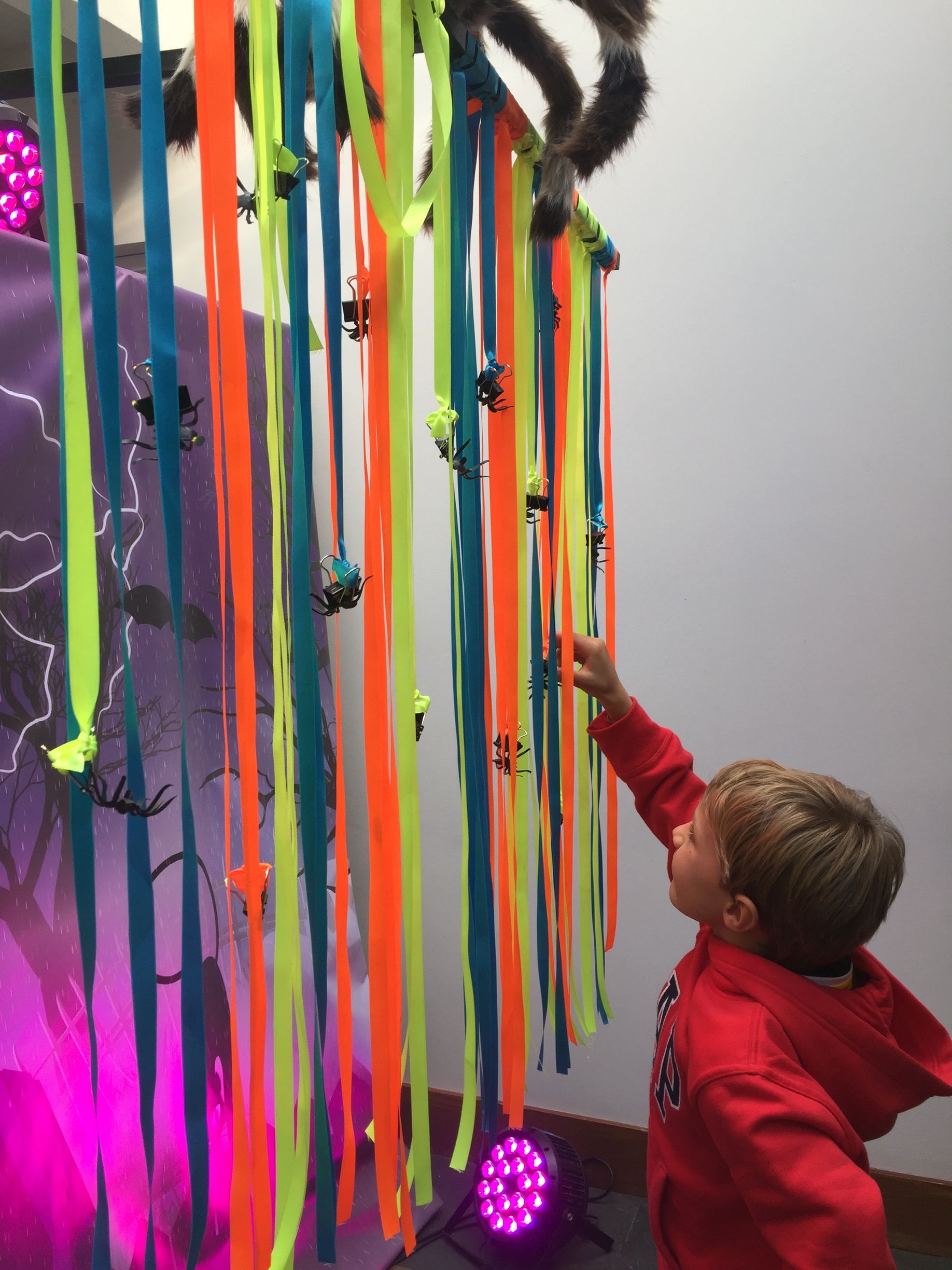 Un niño con una chaqueta roja señala una araña que cuelga de una cinta de colores.