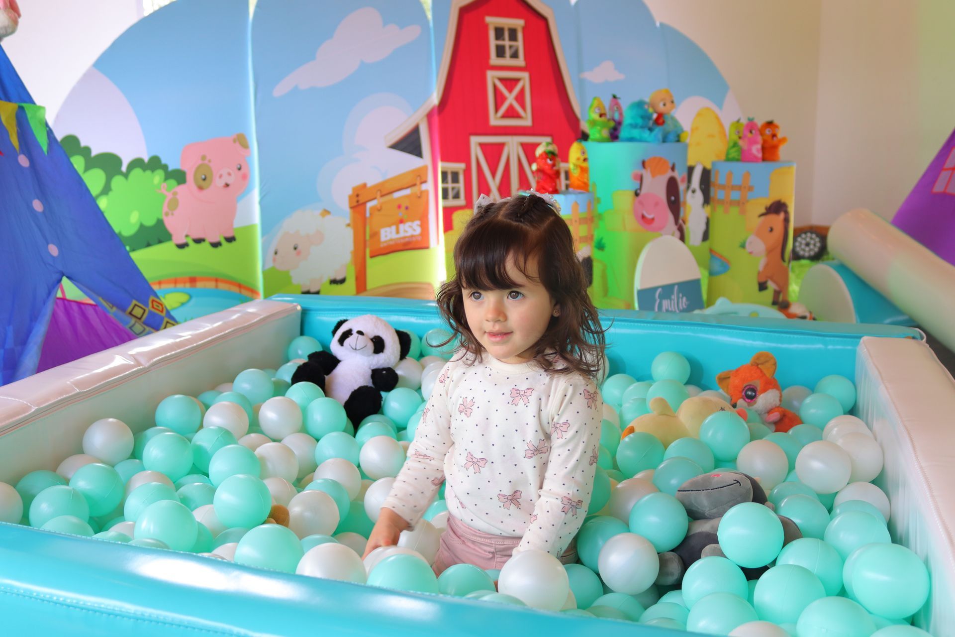 Un niño en una piscina de pelotas de color turquesa, con un juguete de panda y una granja de fondo.
