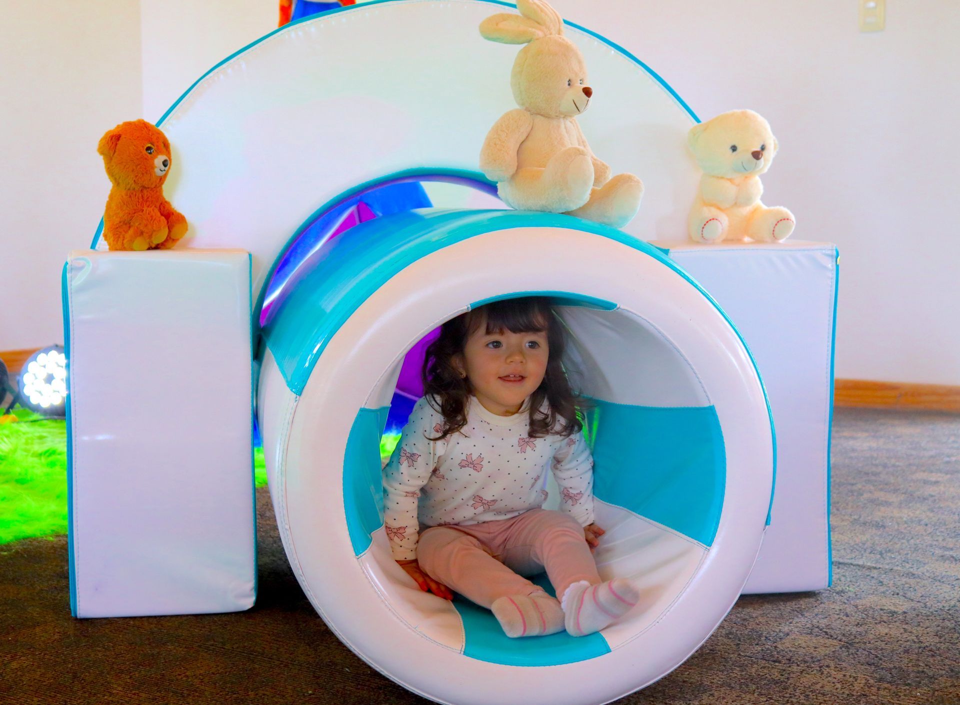 Una niña en un túnel de juegos sonríe, rodeada de animales de peluche y estructuras de juego blancas y azules.