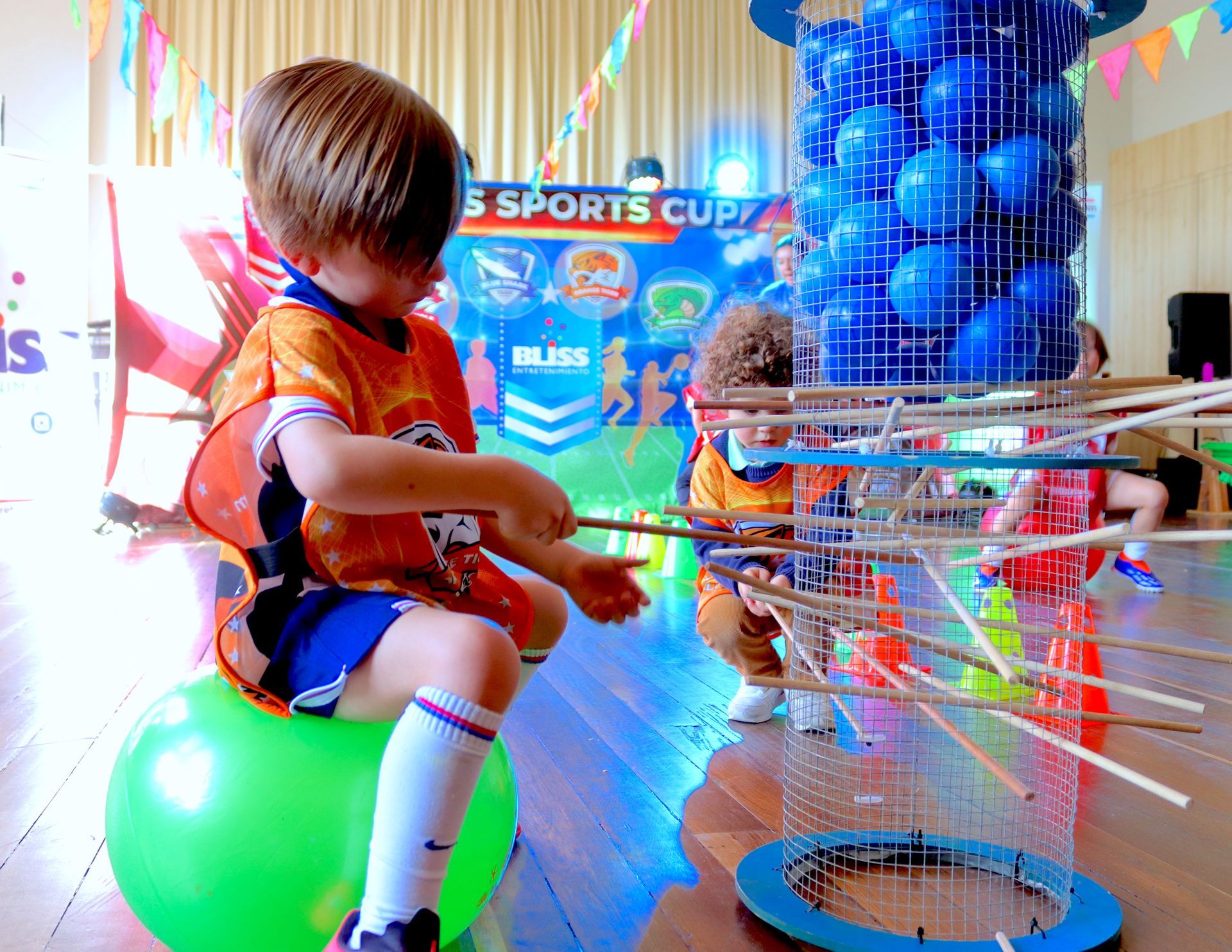 Un niño pequeño está sentado sobre una pelota verde frente a un cartel de una copa deportiva.