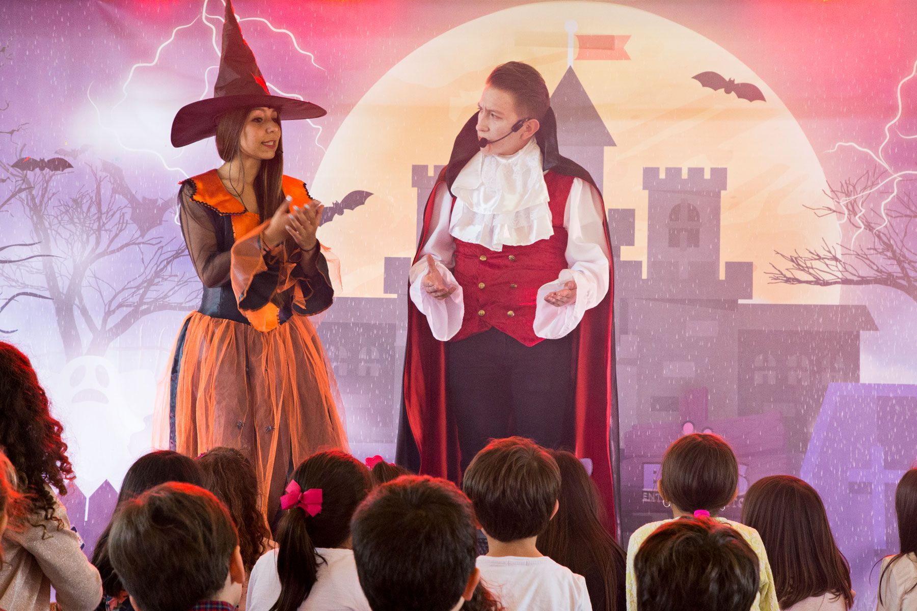 Una bruja y un vampiro están parados en un escenario frente a una multitud de niños.