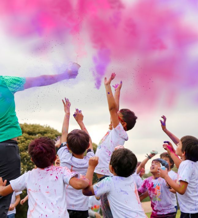 Niños con camisas blancas lanzando polvos de colores al aire durante un vibrante evento al aire libre.