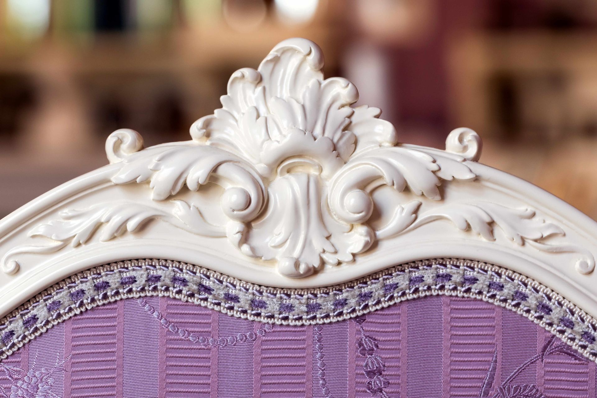 A close-up of a carved white chair back featuring floral details, purple upholstery and braided trim