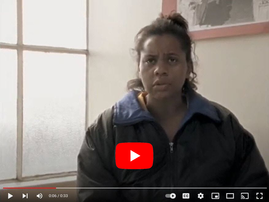 Woman in a dark jacket sits near a window, speaking with a serious expression. A YouTube play button is visible.