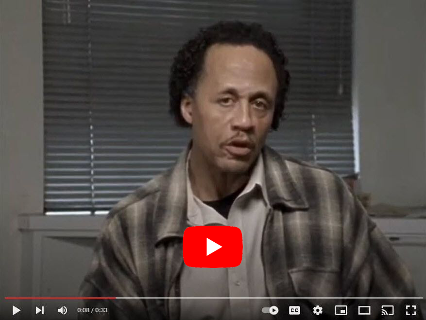 Man in plaid shirt speaking, YouTube video frame. Window blinds in background.