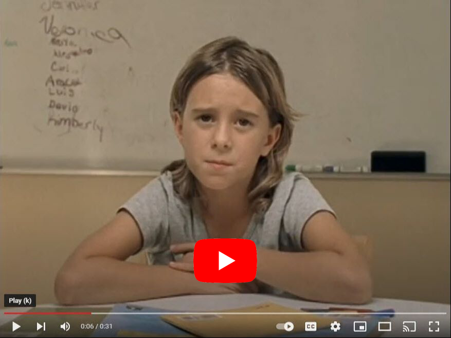 Girl at desk, looking at camera. Whiteboard with names behind her.