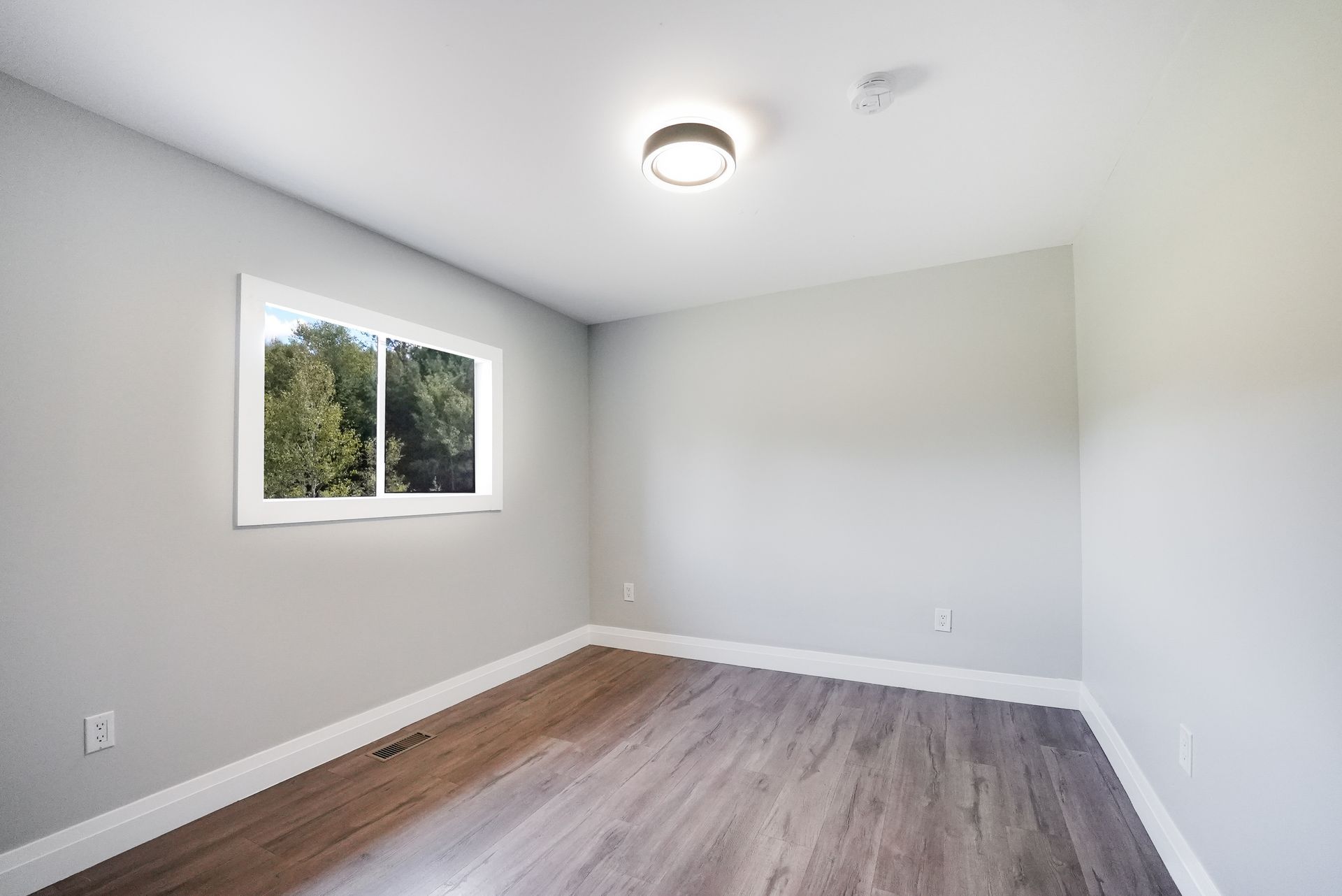 Empty room with gray walls, wooden floor, window, and a ceiling light.
