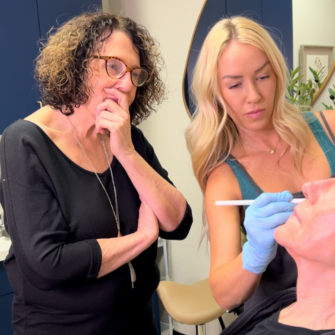 Two women are looking at a man 's face and one of them is wearing blue gloves