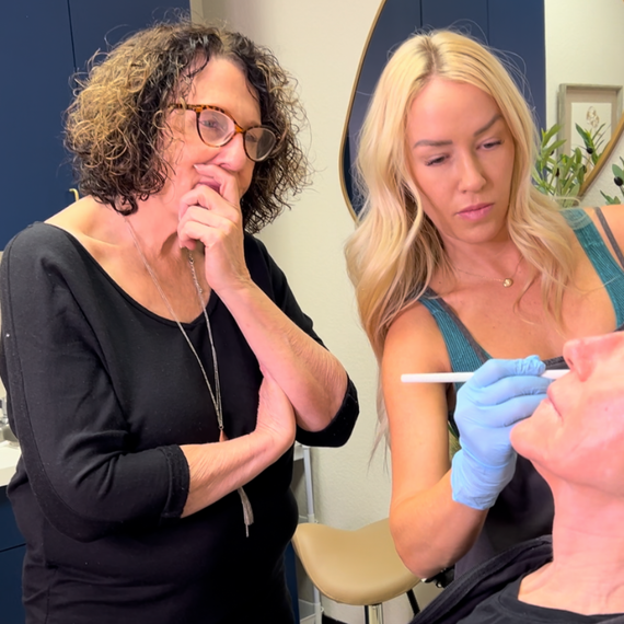 Two women are looking at a man 's face and one of them is wearing blue gloves