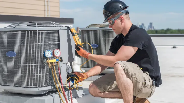 A man is kneeling down on a roof working on an air conditioner.