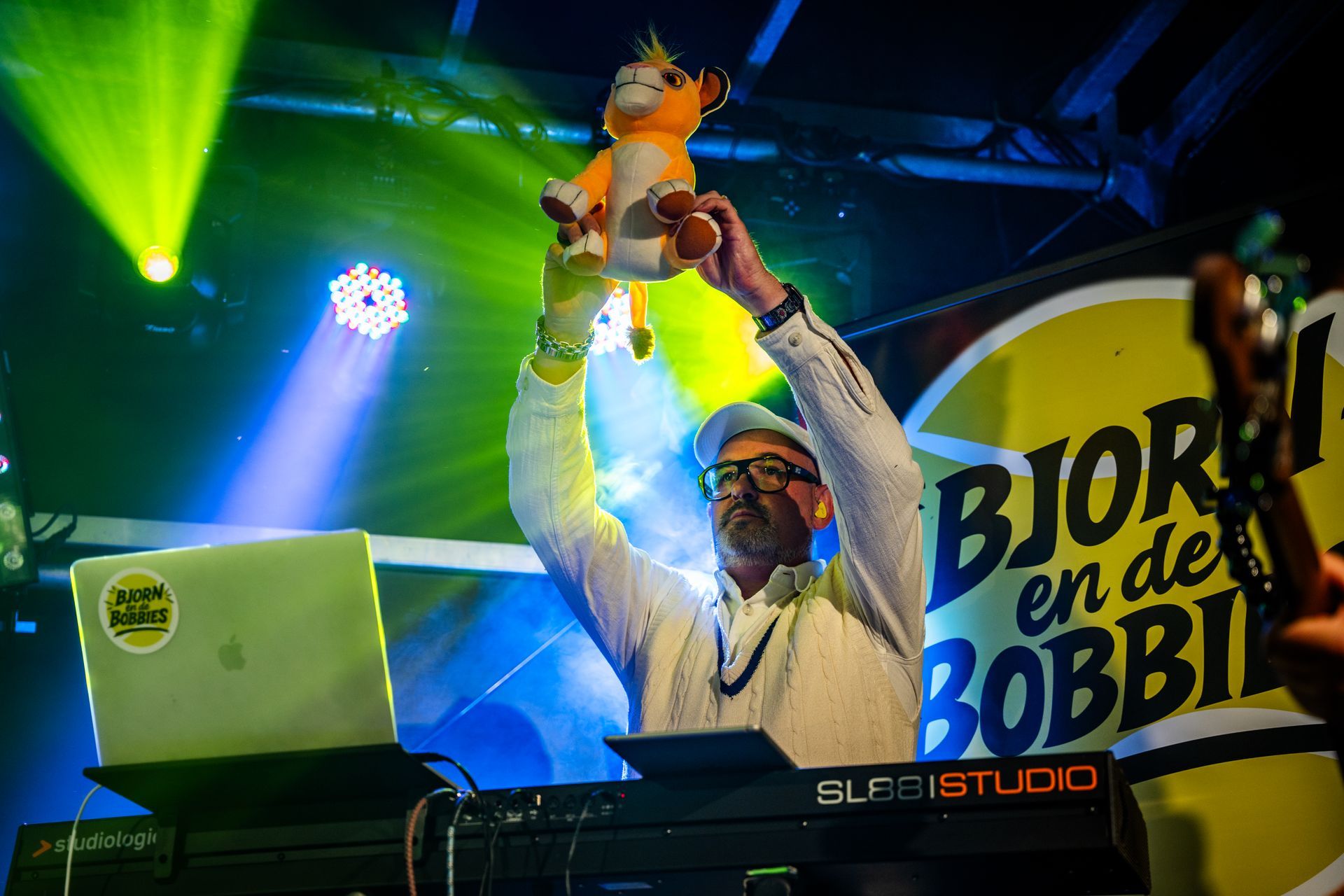 Man op het podium die een knuffeldier omhoog houdt; neonlichten op de achtergrond.