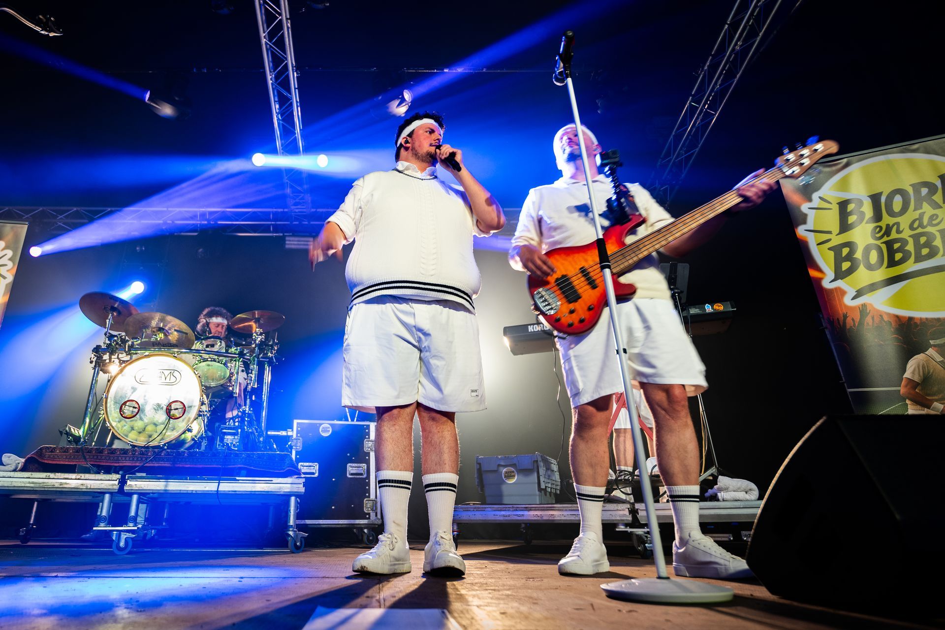 Band treedt op het podium op, gekleed in bijpassende witte sportkleding, met podiumverlichting en drumstel.