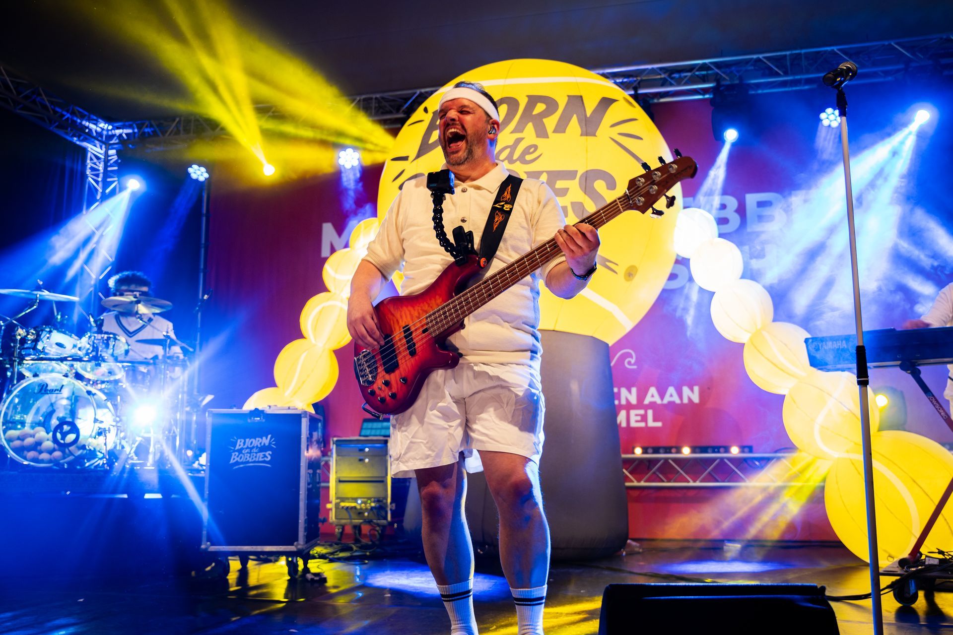 Een man speelt basgitaar op het podium met een band, gekleed in tenniskleding. Het podium is verlicht met blauwe en gele lampen.