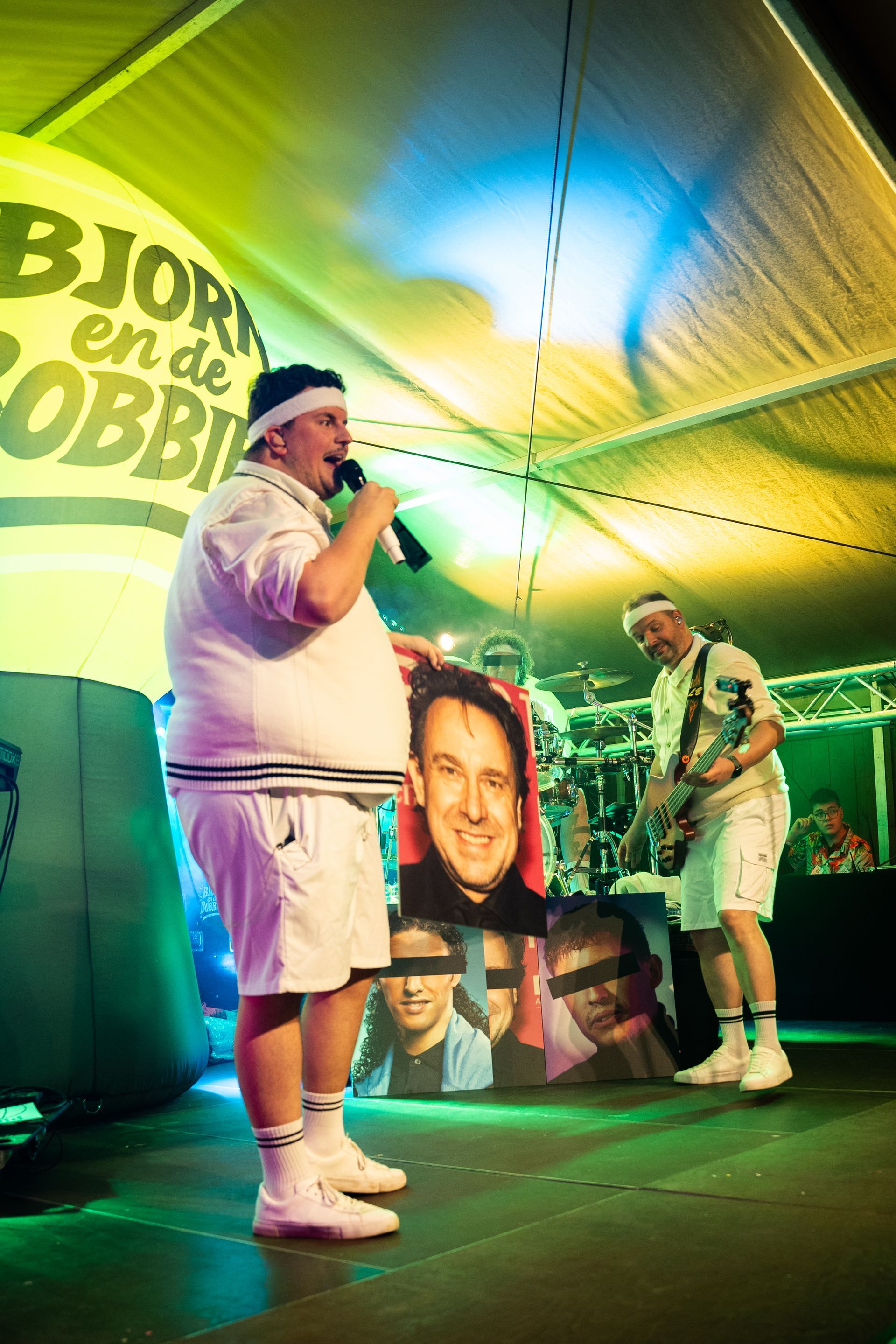 Twee mannen in tenniskleding op het podium; de ene met een microfoon en een portret, de andere met een gitaar.