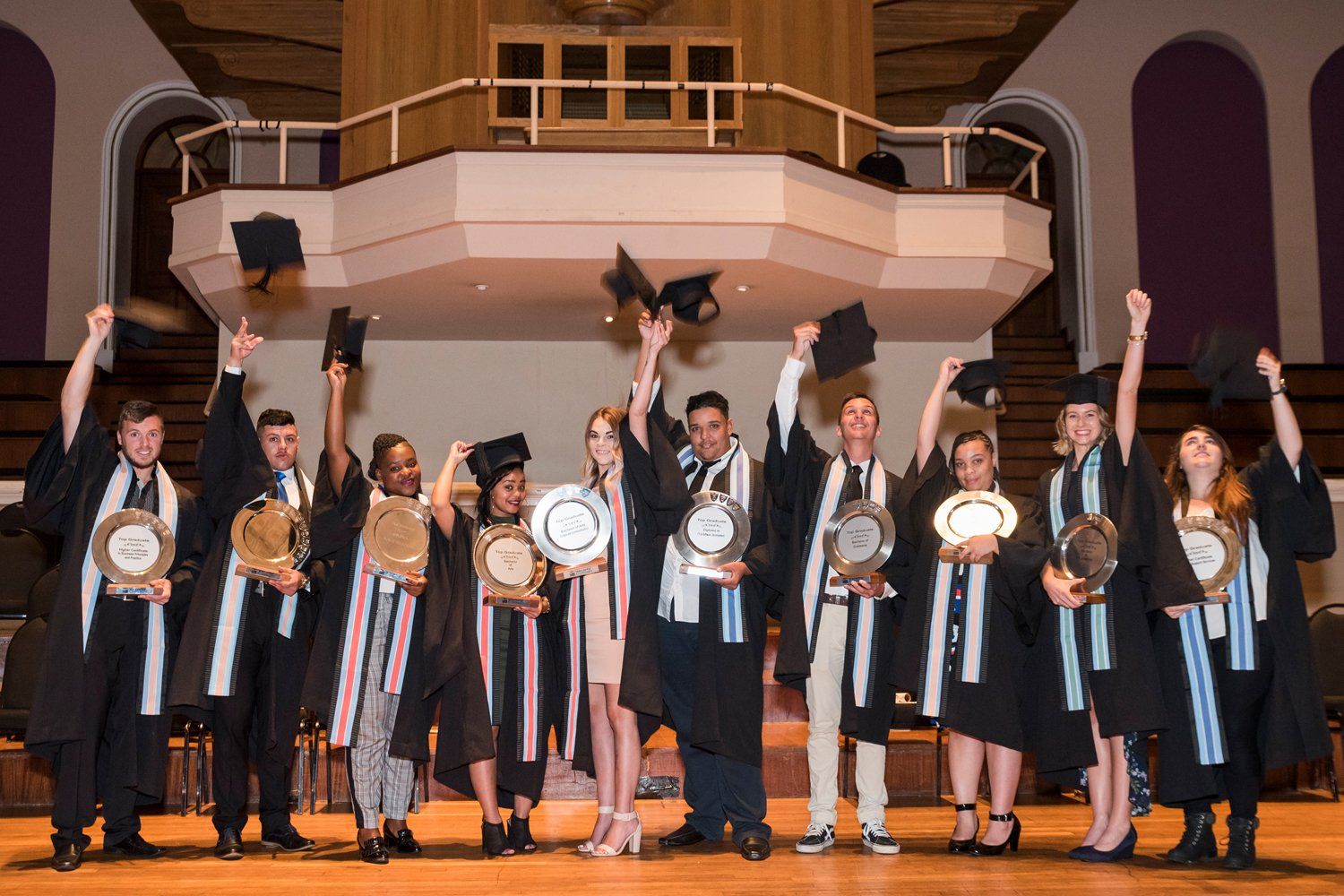 A group of graduates are standing on a stage holding their caps in the air.