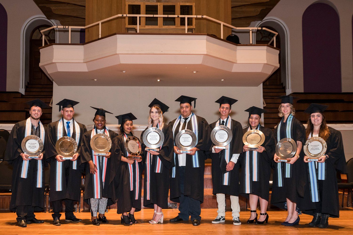 A group of graduates are standing on a stage holding trophies.