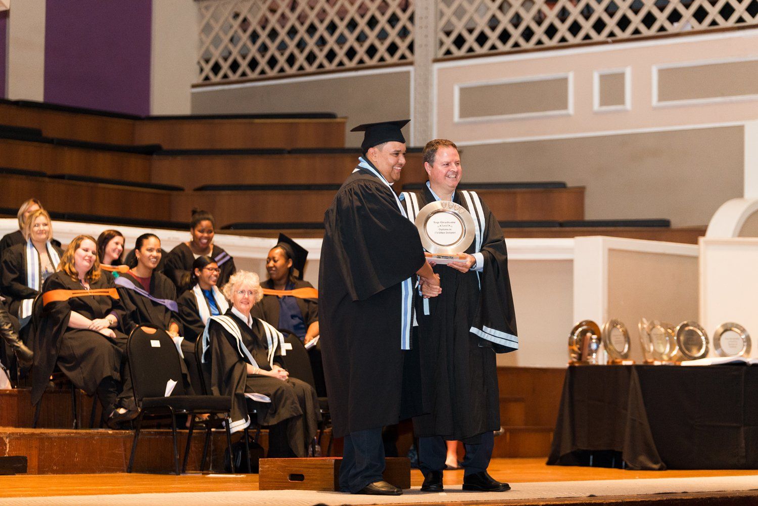 Two men in graduation gowns are standing on a stage holding a plaque.