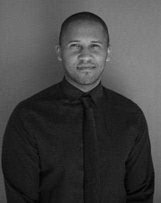 A man in a black shirt and tie is standing in front of a wall.