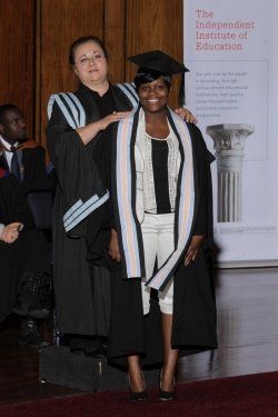 A woman in a graduation cap and gown stands next to another woman