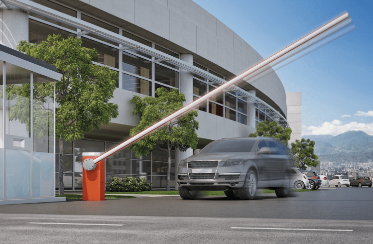 Un automóvil gris circula bajo una puerta de seguridad naranja frente a un edificio moderno.