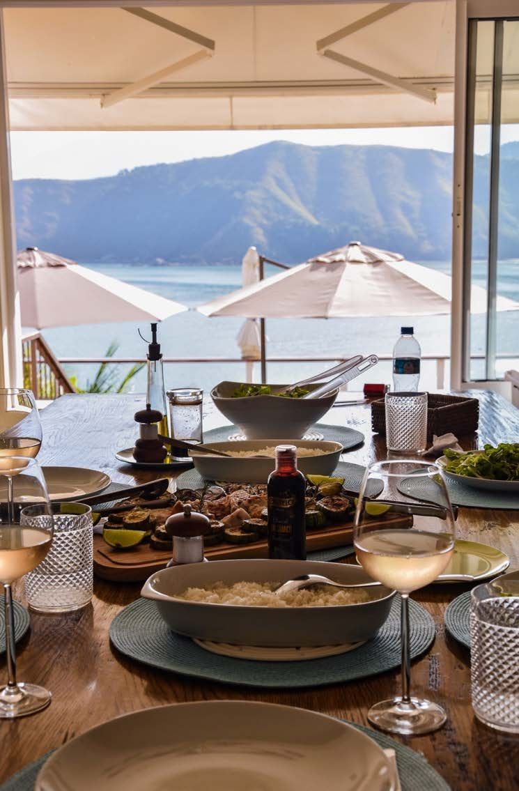 Uma mesa com pratos, tigelas, taças de vinho e vista para o oceano.
