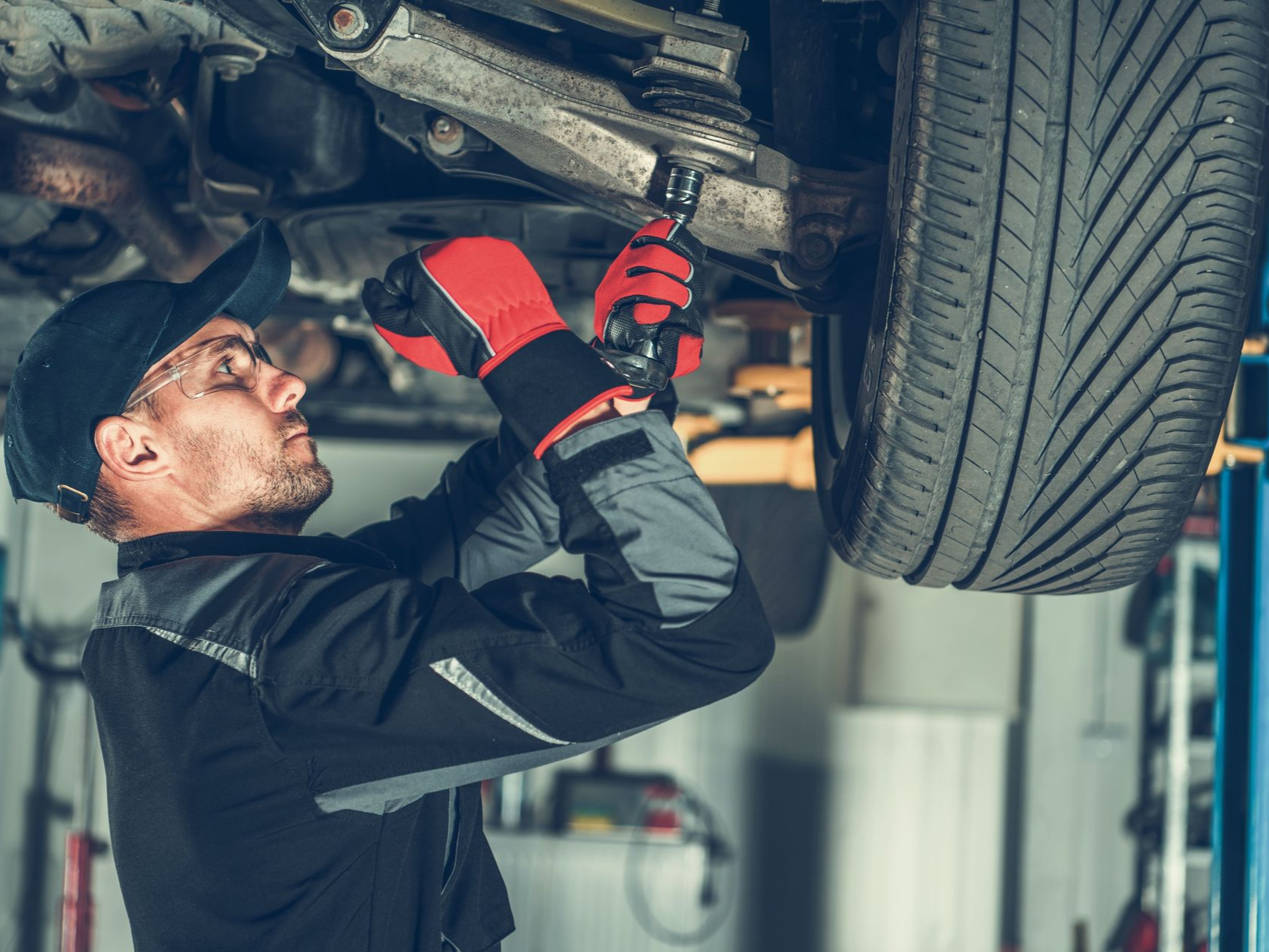 Mechanic in red gloves working on a car's undercarriage in a garage.