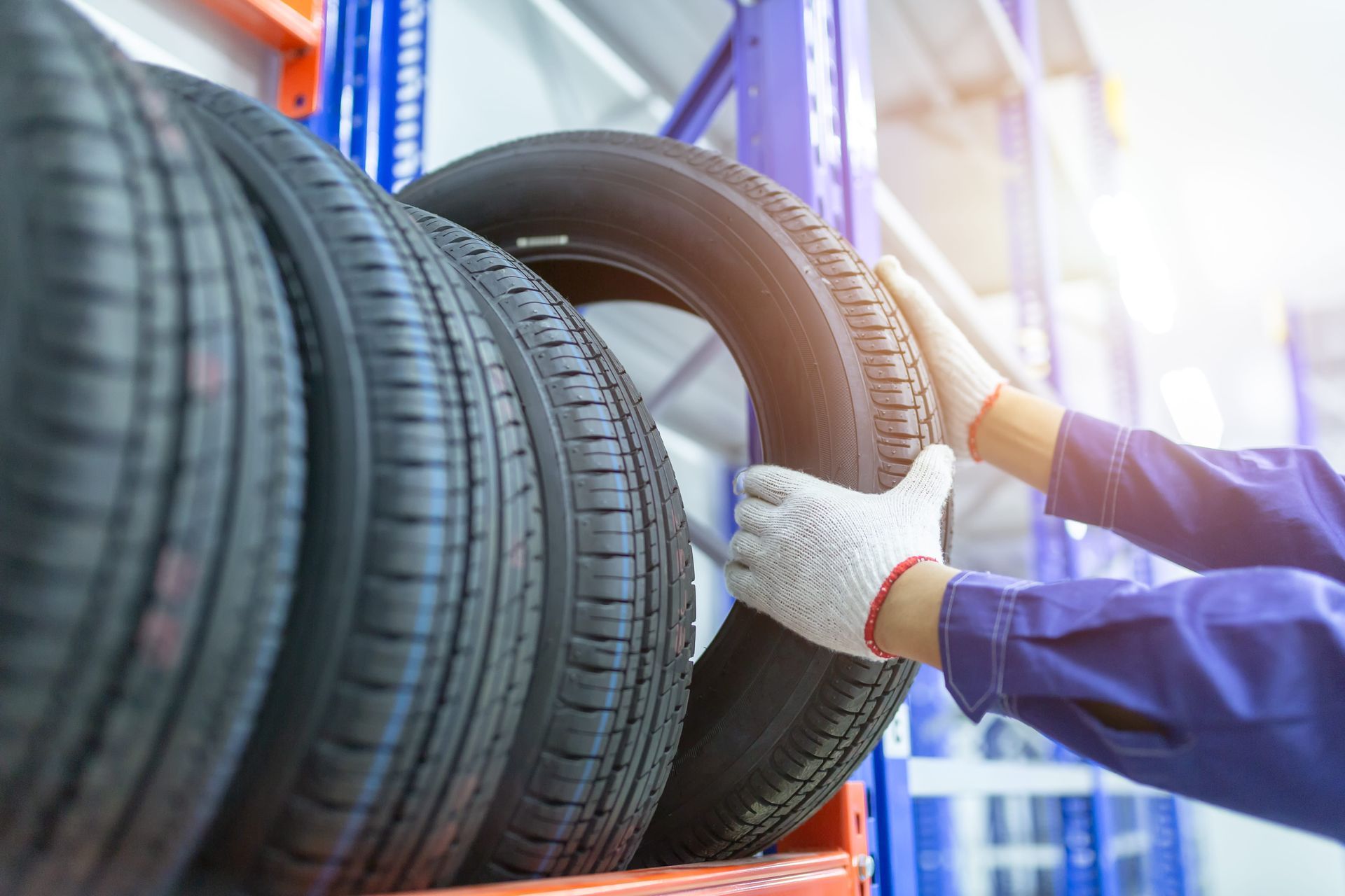 Person in work gloves and blue jumpsuit selecting a tire from a rack in a warehouse.