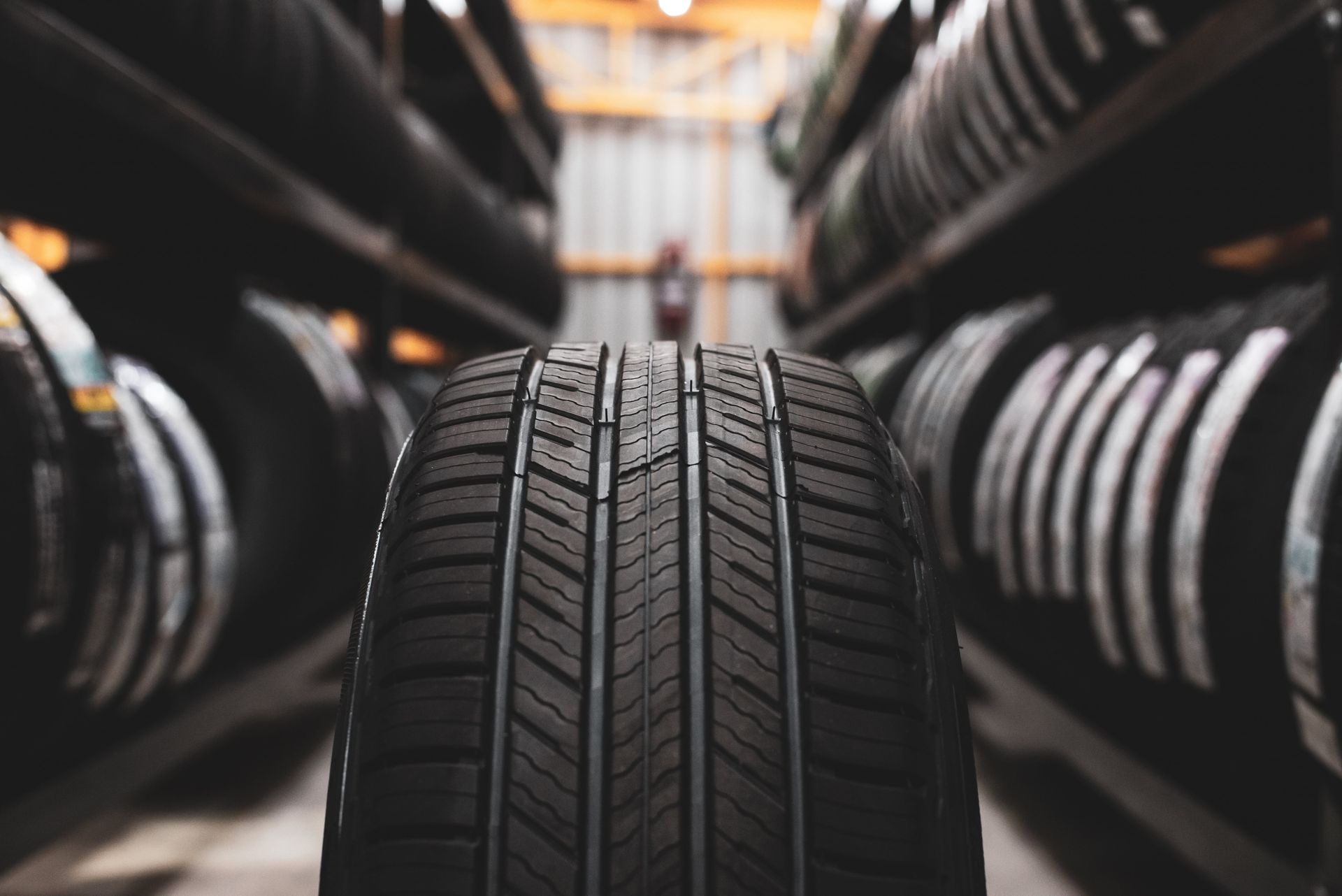 Close-up of a tire with tread, in front of shelves filled with other tires in a tire shop.