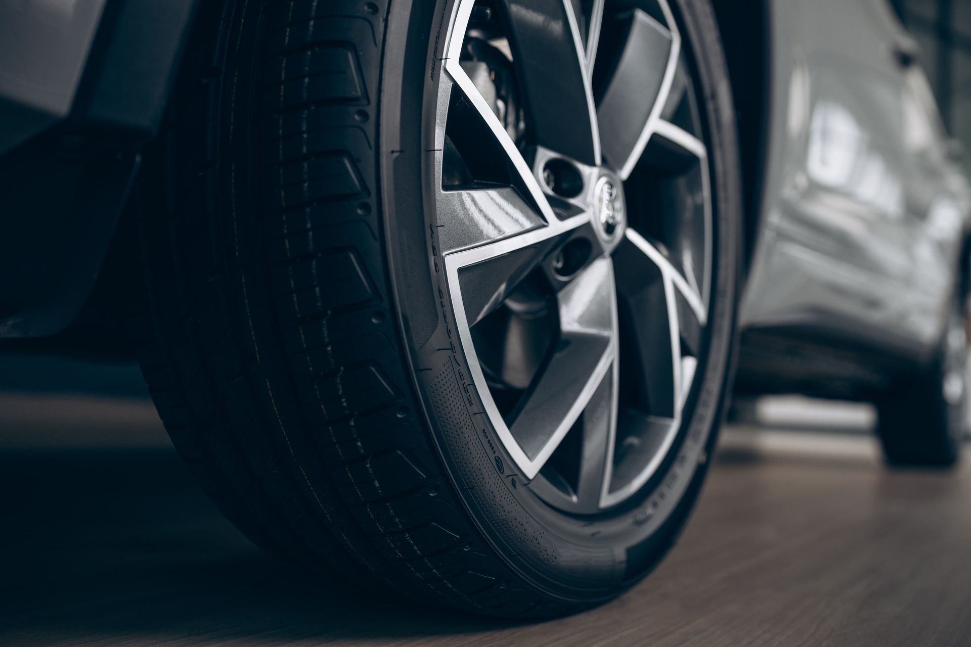 Close-up of a car tire and black and silver rim, on a light brown surface, inside a building.