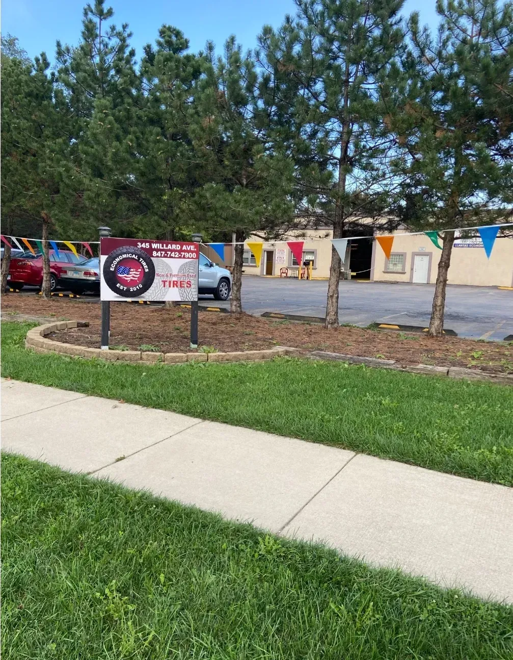 Sign for auto repair shop with cars and building in the background. Green grass and sidewalk in the foreground.