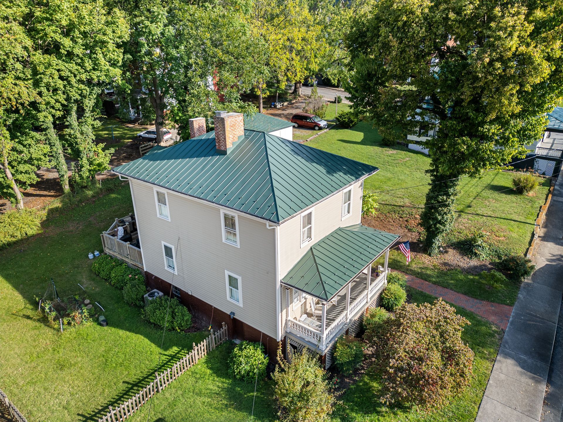 Aerial view of a beige house with a green roof surrounded by trees and a fenced yard