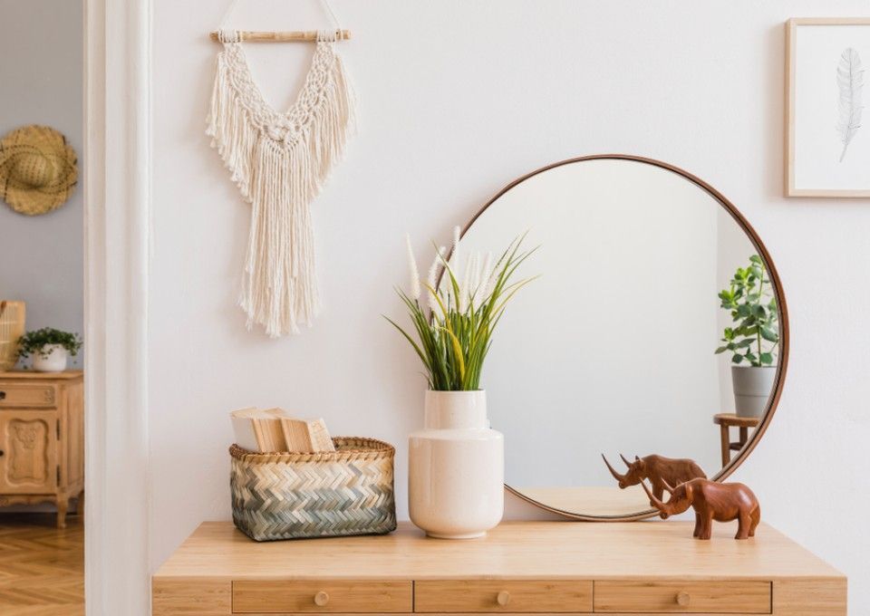 A minimalist entryway with a wooden console, round mirror, macrame wall hanging, woven basket, and rhinoceros figurines.