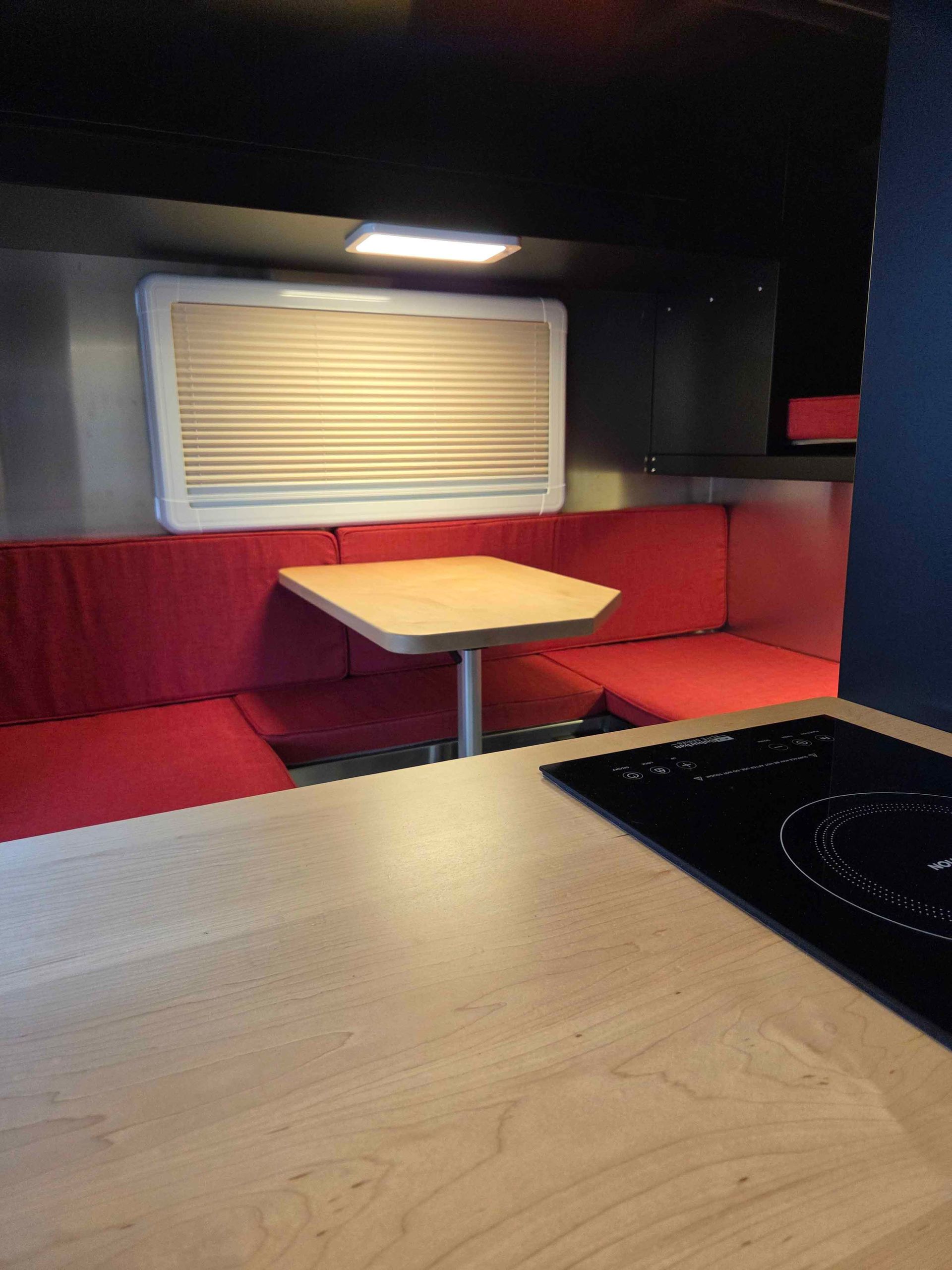 Interior of a camper van: red seating, small table, window, stovetop, and light-colored countertops.