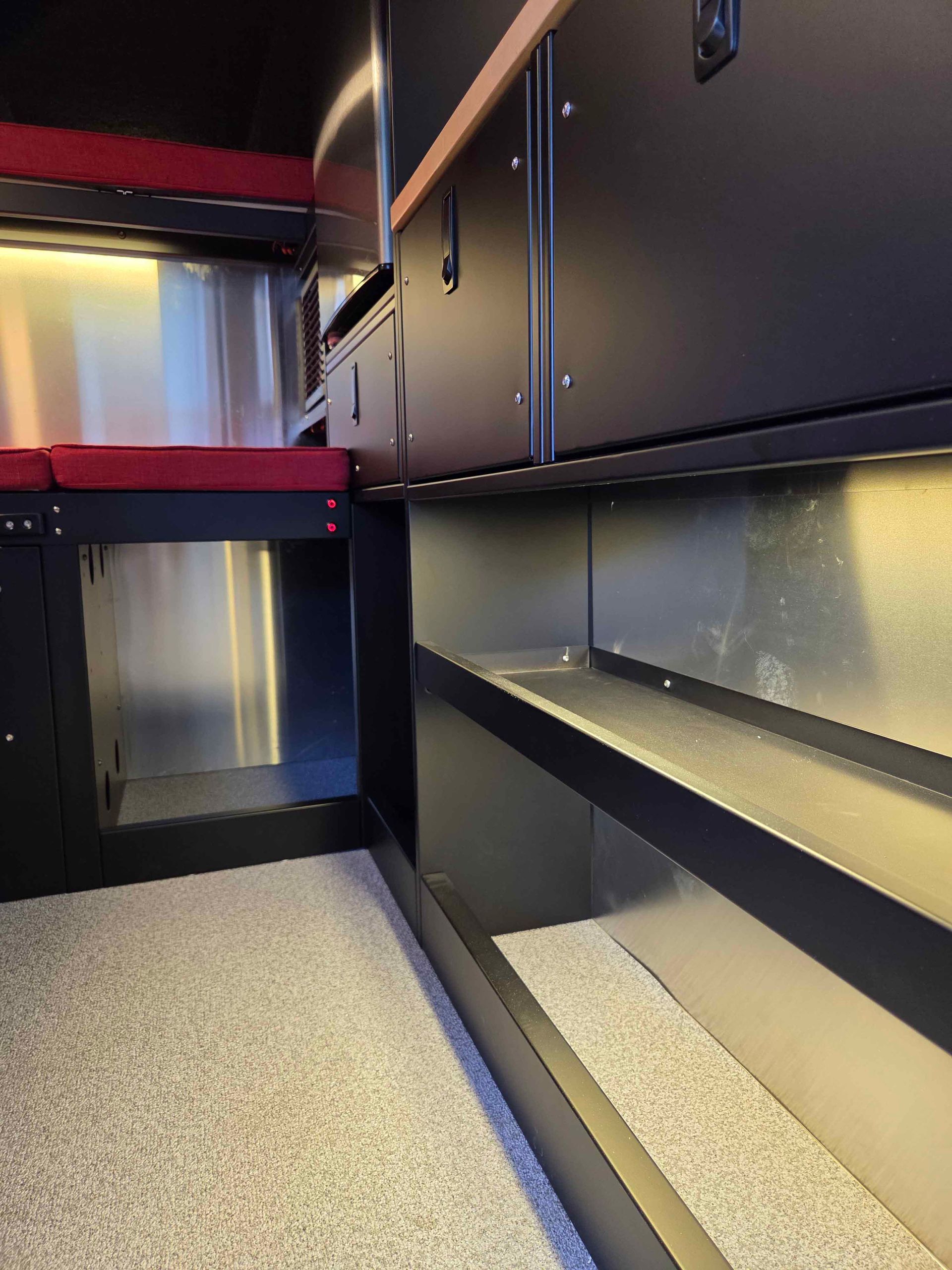 Interior of a black camper with cabinets, red bed, and silver metal walls.
