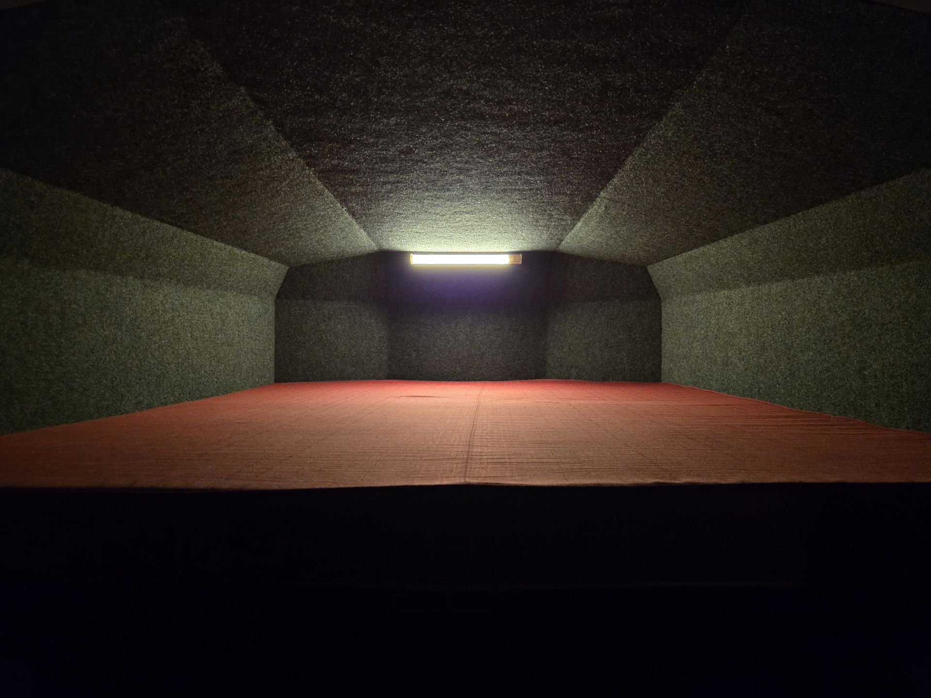 A dark, rectangular room with a red floor and gray walls, illuminated by a strip of light above.