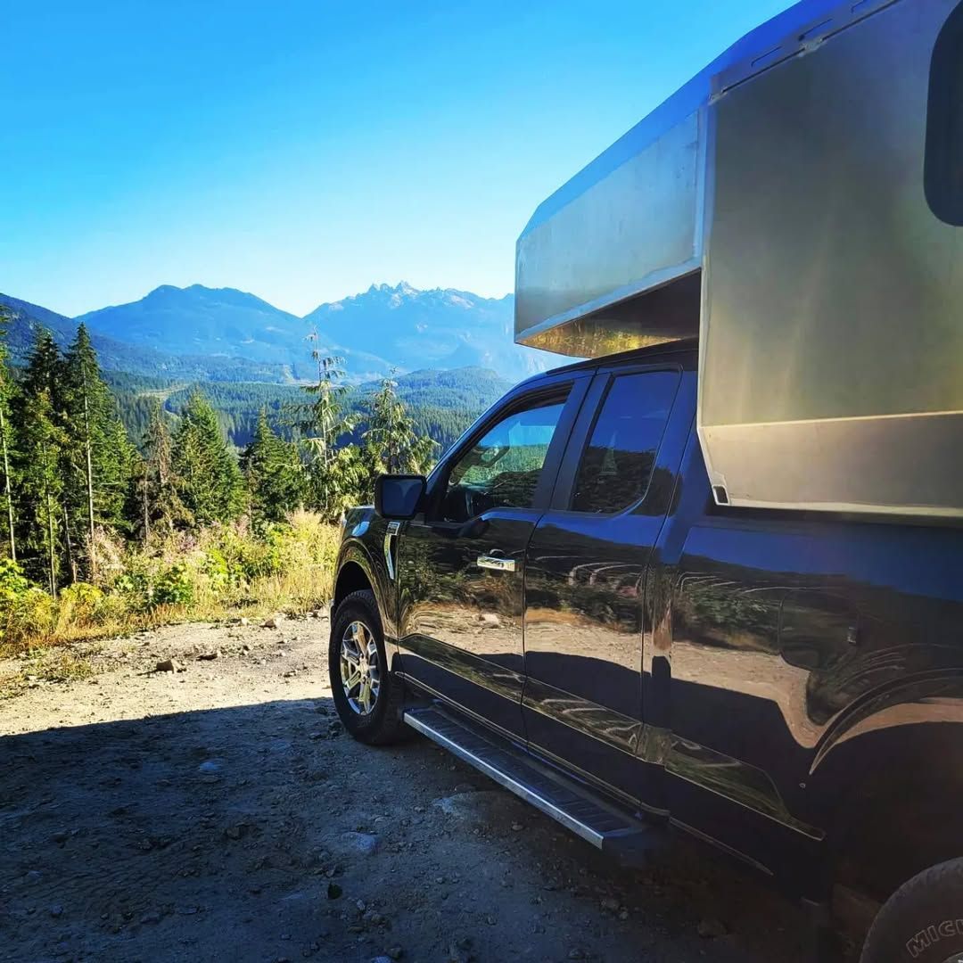 Black pickup truck with camper shell parked on a dirt road overlooking mountains under a blue sky.