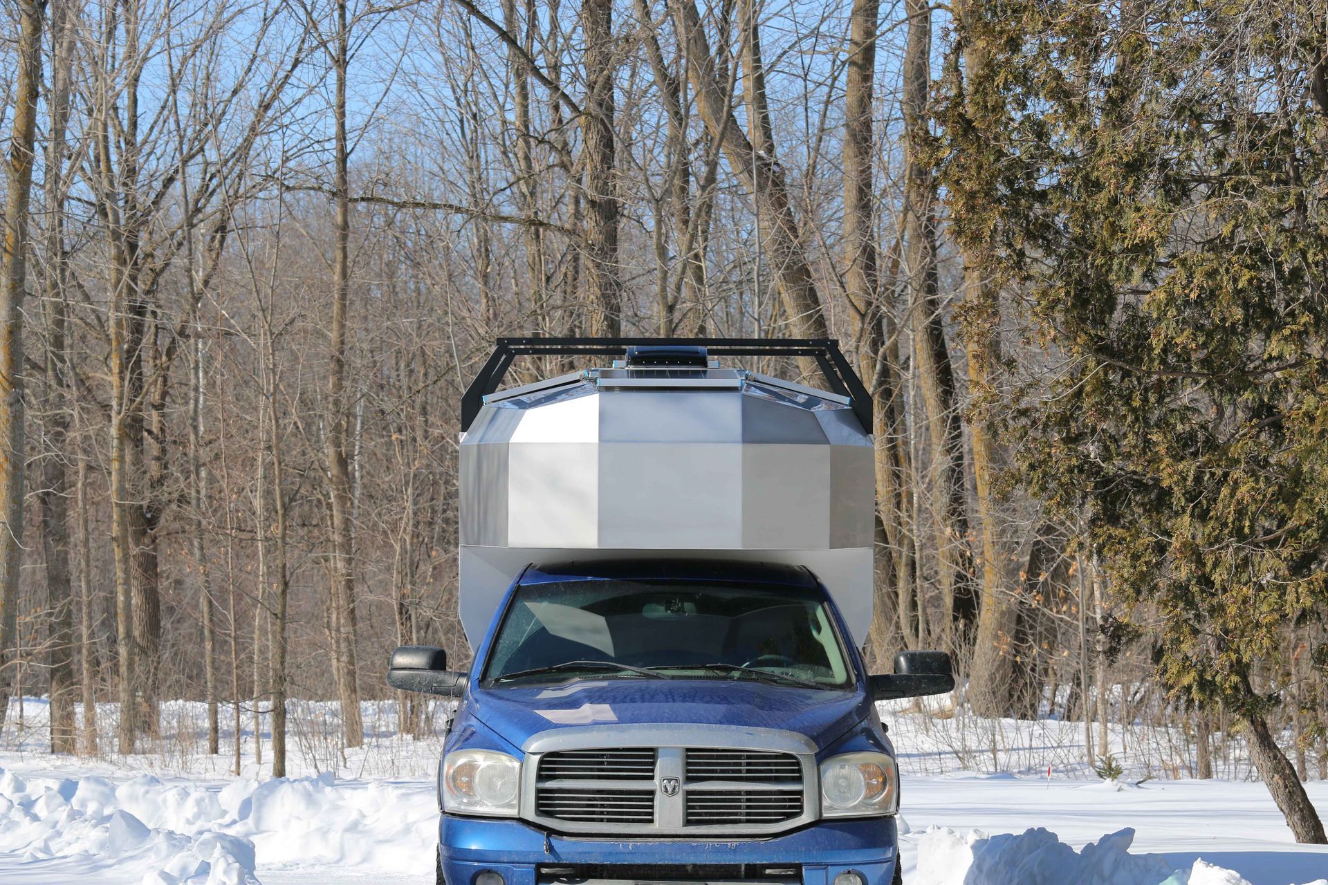 Blue Dodge truck with a silver, geometric camper in a snowy driveway, surrounded by bare trees.