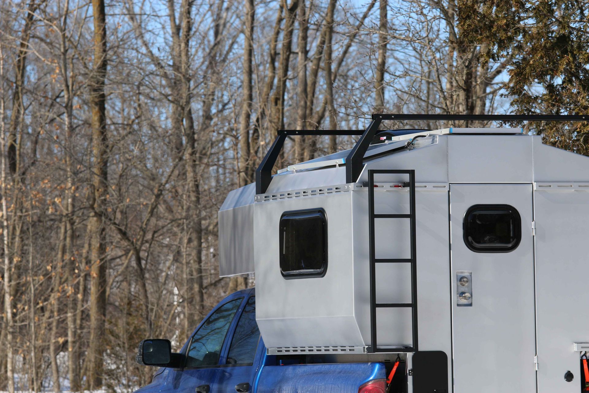 Blue truck with silver camper shell, black ladder and roof rack, in a snowy wooded area.