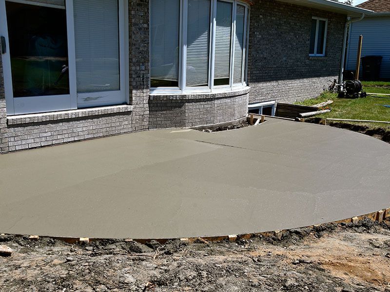 Newly poured concrete patio in front of a brick house, near a bay window and dirt.