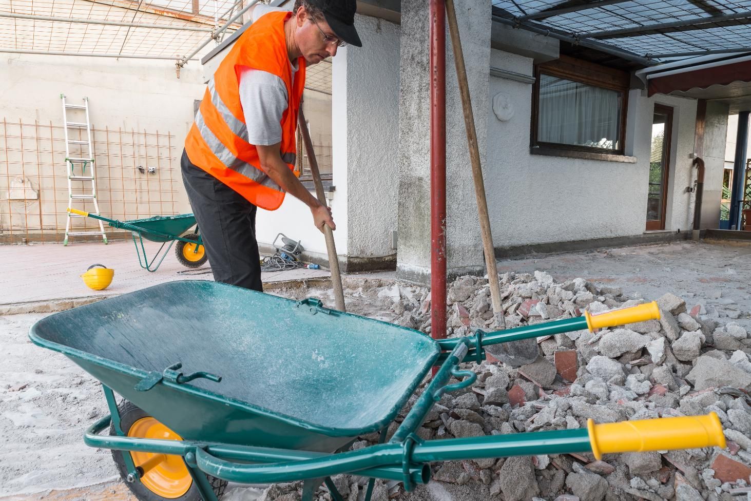 Construction worker using a shovel to load rubble into a green wheelbarrow; orange safety vest, exterior.