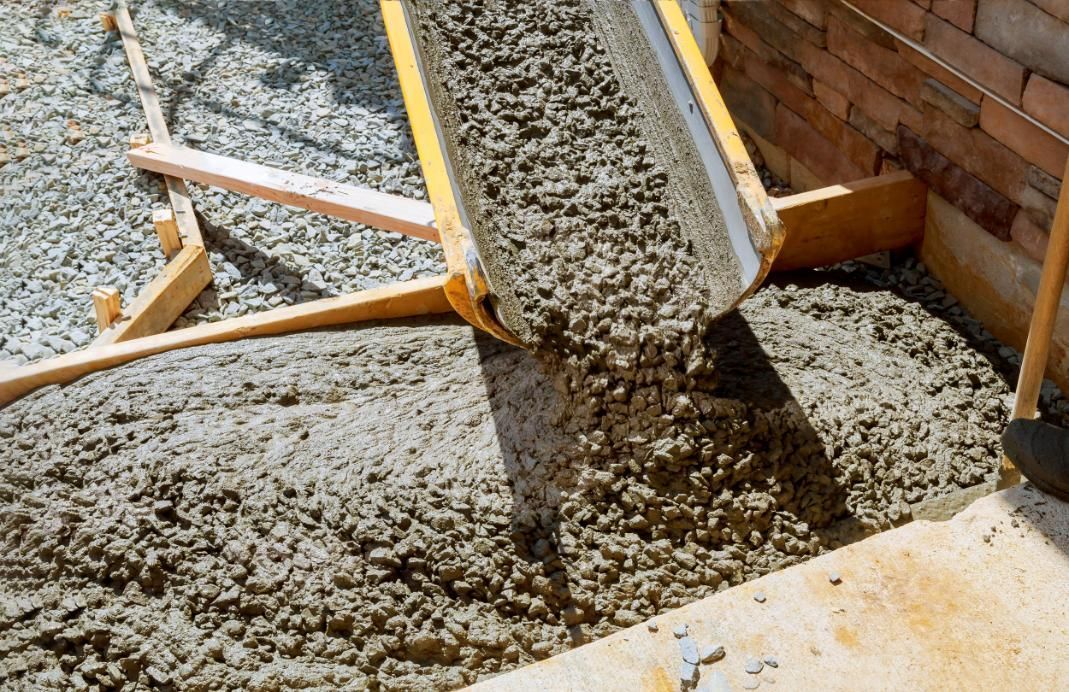 Cement being poured from a yellow chute onto a ground space, next to a brick wall.