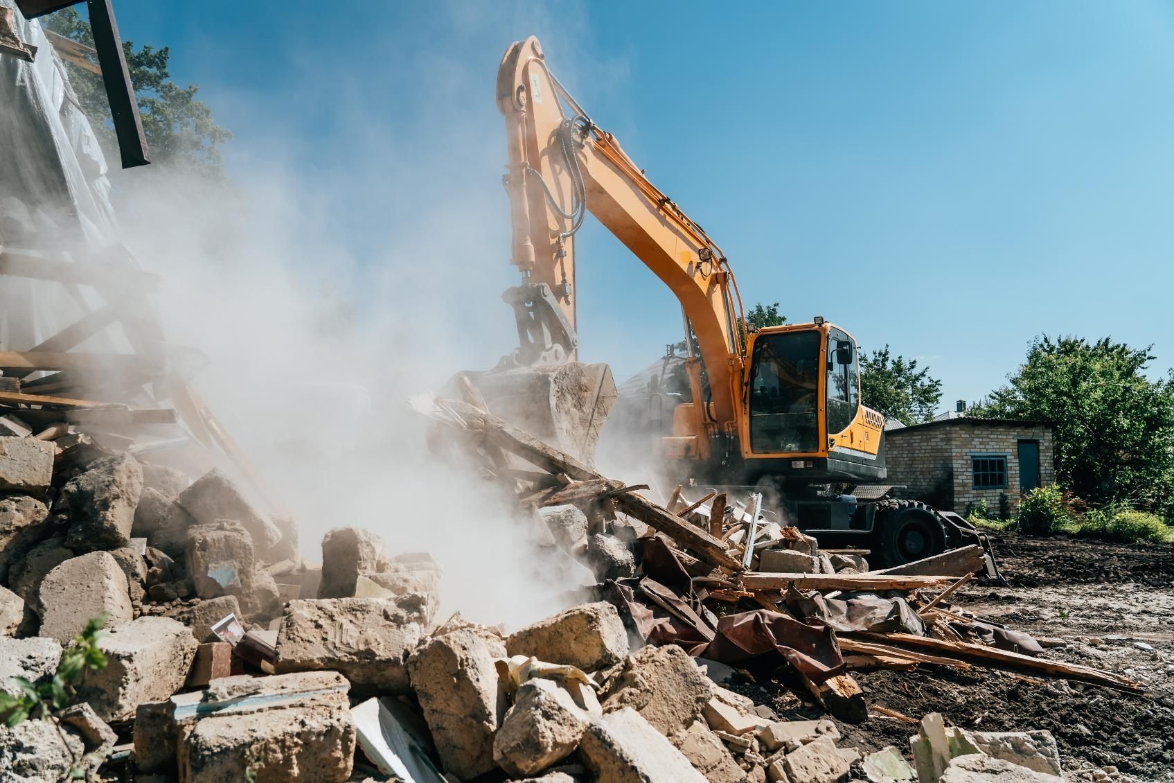 Yellow excavator demolishing a building, raising dust under a clear blue sky.
